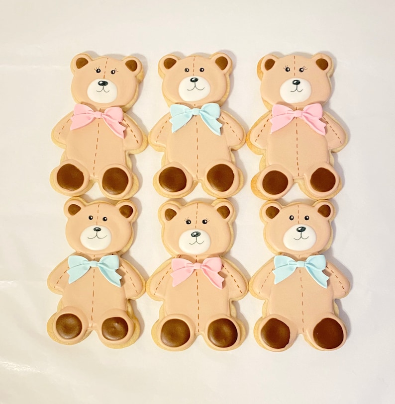 May include: Six decorated sugar cookies shaped like teddy bears. The cookies are brown with white faces and brown eyes. Each cookie has a bow tied around its neck. Three cookies have pink bows and three cookies have blue bows.