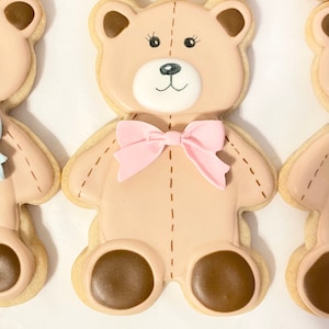 May include: A brown teddy bear cookie decorated with pink icing and a pink bow. The cookie has brown eyes and a brown nose.