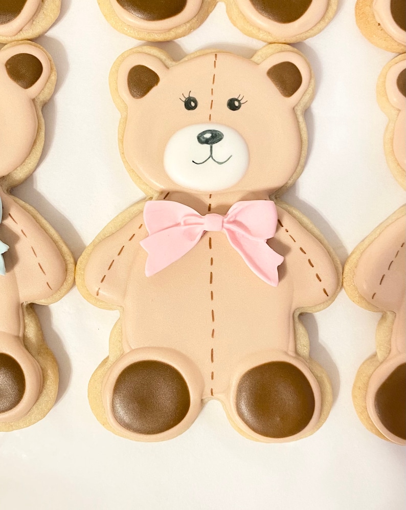 May include: A brown teddy bear cookie decorated with pink icing and a pink bow. The cookie has brown eyes and a brown nose.