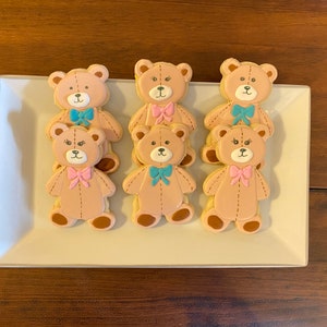 May include: A platter of six decorated sugar cookies shaped like teddy bears. The cookies are decorated with brown, pink, and blue icing. The bears have bows tied around their necks.