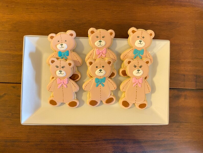 May include: A platter of six decorated sugar cookies shaped like teddy bears. The cookies are decorated with brown, pink, and blue icing. The bears have bows tied around their necks.
