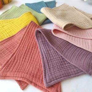 Peut inclure: Un ensemble de huit linges en mousseline de coton de forme triangulaire dans diff&eacute;rentes couleurs, notamment jaune, vert, bleu, beige, rose et violet. Les linges sont doux et textur&eacute;s, avec un motif matelass&eacute; subtil.