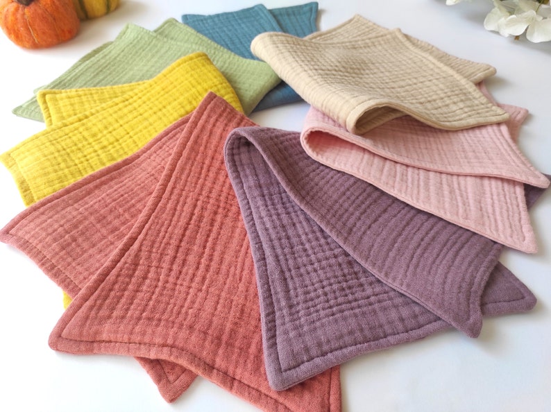 Peut inclure: Un ensemble de huit linges en mousseline de coton de forme triangulaire dans diff&eacute;rentes couleurs, notamment jaune, vert, bleu, beige, rose et violet. Les linges sont doux et textur&eacute;s, avec un motif matelass&eacute; subtil.