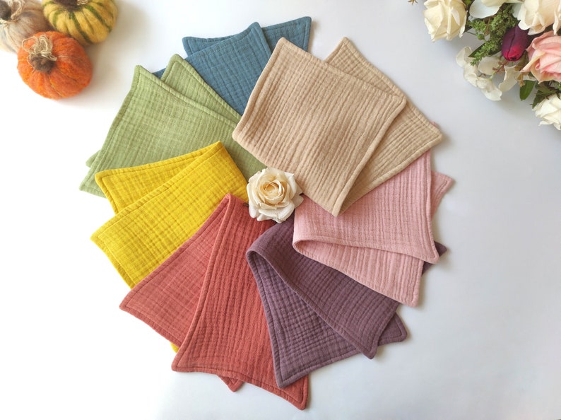 Peut inclure: Un ensemble de huit langes carr&eacute;s en tissu de diff&eacute;rentes couleurs, notamment vert, jaune, orange, rose et violet. Les langes sont dispos&eacute;s en cercle sur une surface blanche.
