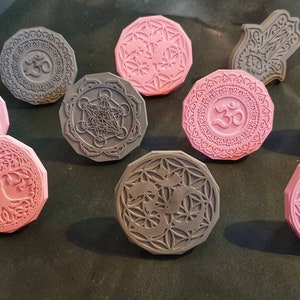 Könnte beinhalten: Eine Sammlung grauer und rosa Gummistempel mit verschiedenen spirituellen und geometrischen Designs. Die Stempel zeigen Symbole wie das Om-Symbol, Metatrons Würfel und den Baum des Lebens. Die Stempel sind achteckig.
