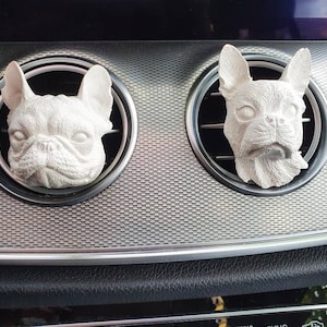 Könnte beinhalten: Zwei weiße französische Bulldoggen-Dekorationen für die Lüftungsschlitze im Auto. Die Hundeköpfe sind geformt und passen in die runden Lüftungsschlitze des Armaturenbretts eines Autos. Das Armaturenbrett hat ein silbernes und schwarzes Design.