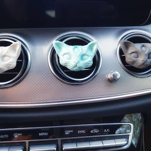 Könnte beinhalten: Drei katzenförmige Auto-Lufterfrischer in Weiß, Hellblau und Grau, jeweils in einer Lüftungsöffnung platziert. Die Katzenköpfe haben drei Augen und befinden sich auf einem silbernen Armaturenbrett.