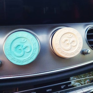 Könnte beinhalten: Zwei Auto-Lufterfrischer mit Om-Symbol. Einer ist hellblau, der andere cremefarben. Beide sind rund und liegen auf einem silbernen Armaturenbrett.