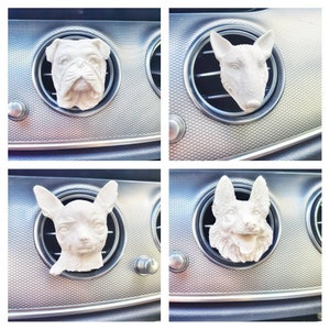 Könnte beinhalten: Vier weiße Auto-Lufterfrischer in Form von Hundeköpfen. Zu den Rassen gehören eine Bulldogge, ein Bullterrier, ein Chihuahua und ein Deutscher Schäferhund. Jeder Hundekopf wird in eine Auto-Luftdüse eingesetzt.