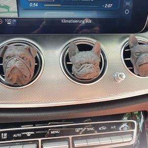 Könnte beinhalten: Drei braune, hundeförmige Auto-Luftauslassdekorationen. Die Hunde sind eine Bulldogge und zwei französische Bulldoggen. Die Dekorationen sind in den Luftauslässen eines Armaturenbretts angebracht. Das Armaturenbrett ist silber und schwarz.