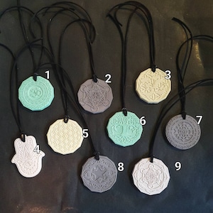Könnte beinhalten: Eine Sammlung von neun Anhänger-Halsketten mit schwarzen Kordeln. Die Anhänger sind in verschiedenen Farben erhältlich, darunter Mintgrün, Grau und Off-White. Jeder Anhänger zeigt ein anderes geometrisches oder spirituelles Design, wie ein Om-Symbol, einen Metatron-Würfel und einen Lebensbaum.