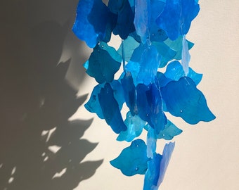 May include: A hanging mobile featuring layered, translucent blue petal-shaped elements. The petals vary in shades of blue, creating a gradient effect. The mobile casts a shadow on a white wall.