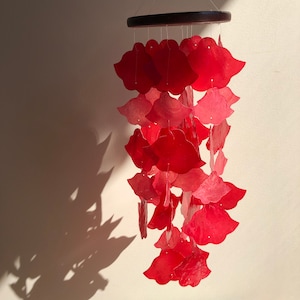 May include: A red wind chime with petal-shaped pieces suspended from a dark wooden ring. The petals are in varying shades of red. The chime hangs from thin strings. The overall design is decorative.