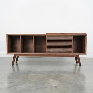 May include: A mid-century style wooden media console in a warm brown finish. It has three open compartments, a sliding door with horizontal slats, and angled legs. The console is designed for storage and display.