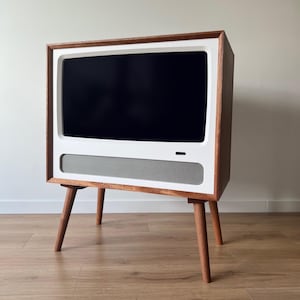 Puede incluir: Un mueble de televisión de estilo retro con un marco blanco y una pantalla oscura. El mueble tiene un exterior de madera y está sostenido por cuatro patas de madera en ángulo. Una rejilla de altavoz es visible debajo de la pantalla.