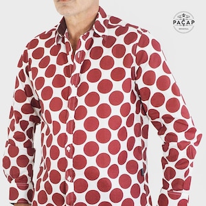 Peut inclure: Une chemise boutonnée à manches longues blanche avec un motif de pois rouges. La chemise a un col et une fermeture boutonnée. Le nom de la marque "PACAP ORIGINAL" est visible sur la chemise.