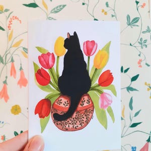 May include: A watercolour illustration of a black cat perched on a decorative vase filled with red, yellow, and pink tulips. The vase features a detailed floral pattern. The artwork is on a white card.