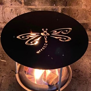 May include: A black metal fire pit cover with a cut-out design of a dragonfly. The fire pit is lit and the flames are visible through the dragonfly design.