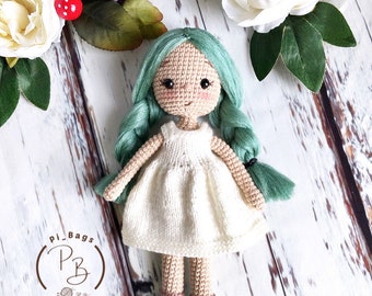Könnte beinhalten: Handgemachte Häkelpuppe mit mintgrünen Haaren und einem weißen Kleid. Die Puppe hat rosige Wangen und blaue Schuhe. Die Puppe ist von weißen Blumen und Grün umgeben. Die Puppe ist etwa 18 cm groß.