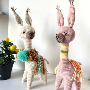 May include: Two crocheted llama toys, one in beige with a brown and teal pom-pom scarf and the other in pink with a multi-colored scarf.
