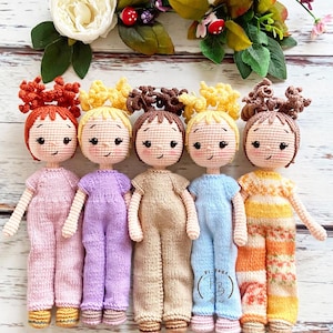 以下が含まれることがあります： 異なる色の服を着た、4体の編みぐるみ人形。人形は茶色、黄色、赤色の髪をしています。人形はピンク、紫、ベージュ、黄色とオレンジのストライプの服を着ています。人形は白い木製の表面に座っています。