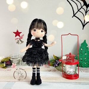 K&ouml;nnte beinhalten: Eine geh&auml;kelte Puppe, die ein schwarzes Kleid mit wei&szlig;em Kragen und schwarz-wei&szlig;en gestreiften Socken tr&auml;gt. Die Puppe hat schwarze Haare und braune Augen. Die Puppe steht auf einer Holzoberfl&auml;che mit einer roten Laterne und einem gr&uuml;nen Weihnachtsbaum im Hintergrund.