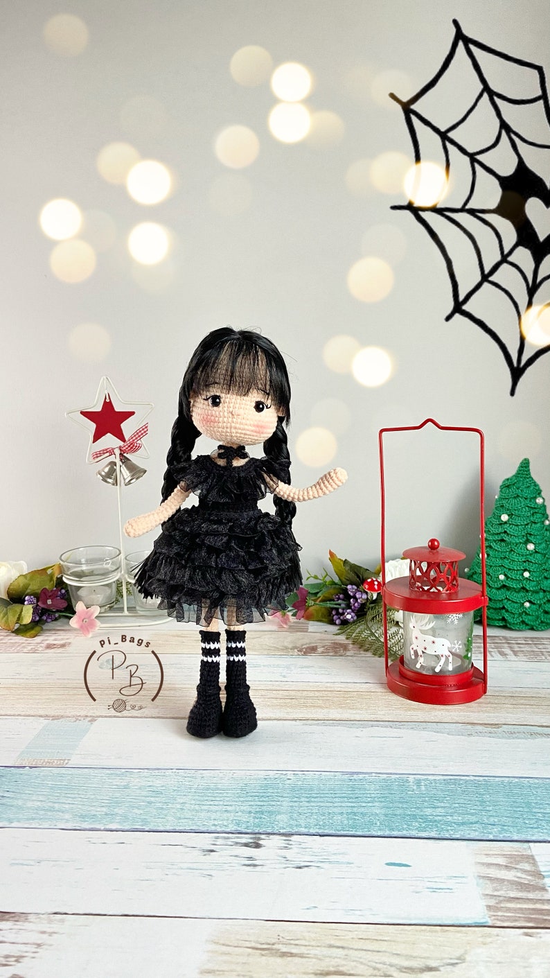 K&ouml;nnte beinhalten: Eine geh&auml;kelte Puppe, die ein schwarzes Kleid mit wei&szlig;em Kragen und schwarz-wei&szlig;en gestreiften Socken tr&auml;gt. Die Puppe hat schwarze Haare und braune Augen. Die Puppe steht auf einer Holzoberfl&auml;che mit einer roten Laterne und einem gr&uuml;nen Weihnachtsbaum im Hintergrund.