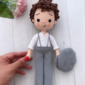 May include: A crocheted doll with brown hair, a white shirt, gray pants, and a gray hat. The doll is standing on a white wooden surface.