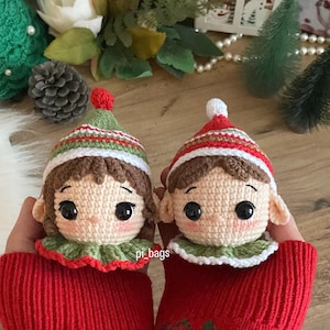May include: Two crocheted elf ornaments with brown hair, green and white hats, and red and white striped trim. The ornaments are wearing green and white ruffled collars and are held in a pair of red knitted mittens with green and white accents. The text "pi_bags" is visible on the mittens.