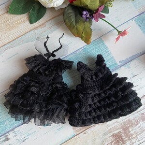 K&ouml;nnte beinhalten: Zwei schwarze Puppenkleider, eines aus Spitze und T&uuml;ll, das andere geh&auml;kelt, beide mit mehreren R&uuml;schen.