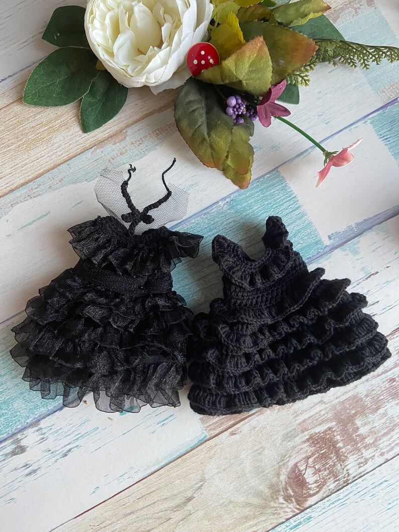 K&ouml;nnte beinhalten: Zwei schwarze Puppenkleider, eines aus Spitze und T&uuml;ll, das andere geh&auml;kelt, beide mit mehreren R&uuml;schen.