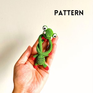 May include: A green crocheted frog with large, googly eyes and a red bow tie. The frog has red feet and black stripes on its body. The word "PATTERN" is written in black, block letters above the frog.