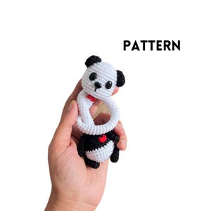 May include: A handmade crochet panda bear toy with a white body, black accents, and a red bow tie. The panda is attached to a white ring. The word "PATTERN" is displayed in black text.