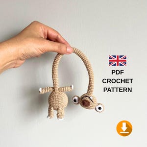 Pode incluir: Um brinquedo de crochê bege com corpo redondo, membros pequenos e um pescoço longo e curvo que leva a uma cabeça com dois olhos brancos grandes e um nariz marrom. O texto PDF CROCHET PATTERN é exibido com um ícone de download.