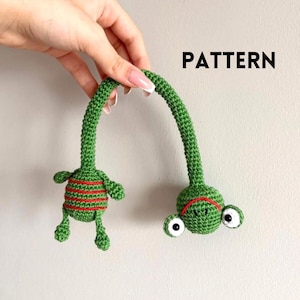 May include: A green crocheted alien toy with red stripes and large white eyes. The toy has long, curved arms and legs. The word "PATTERN" is printed in black, uppercase letters.