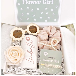 May include: A gift box with a floral theme, containing a white tumbler with a name, flower-shaped sunglasses, a rose, a scrunchie, a pearl hair clip, and a card that says "Will You Be My Flower Girl".