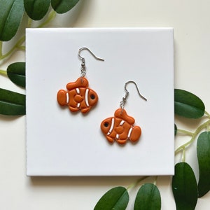 May include: A pair of orange and white clownfish dangle earrings. The earrings are made of polymer clay and have silver hooks.