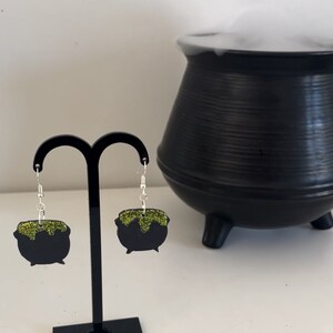 May include: A pair of black cauldron earrings with green glitter accents. The earrings are hanging from a black earring stand. A black cauldron with steam coming out of it is in the background.