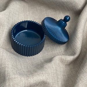 May include: A dark blue, round trinket box with a fluted lid and a small knob on top. The box is sitting on a light brown fabric surface.