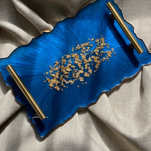 May include: A rectangular, blue resin tray with gold leaf accents and brass handles. The tray has an organic edge and is designed for serving or display. The gold leaf is concentrated in the centre of the tray.