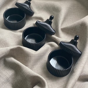 May include: Three black, round containers with lids are displayed on a neutral-toned fabric. The containers feature a ribbed design, and the lids are off, showing the inside. The lids have a decorative, rounded top.