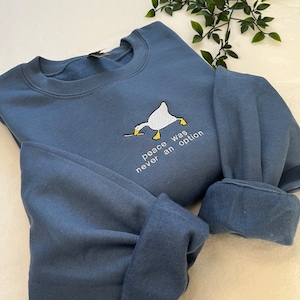 Pode incluir: Uma camisa de frio azul com um ganso branco segurando uma faca e o texto "peace was never an option".