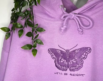 Op de afbeelding: Een lavendelkleurige hoodie met een vlinderprint en de tekst "We'll be alright" geborduurd op de voorkant.