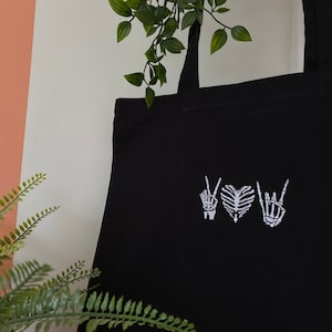 Puede incluir: Bolsa de tela negra con manos de esqueleto bordadas en blanco haciendo señales de paz y una forma de corazón.