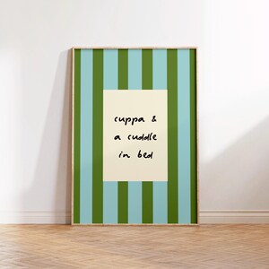 May include: A framed print with a striped background in shades of blue and green. A cream-colored rectangle in the center displays the handwritten text: "cuppa & a cuddle in bed."