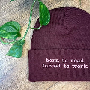 Pode incluir: Um gorro de malha bordô com o texto bordado "born to read forced to work" em branco. O gorro é dobrado na aba. Uma planta verde com folhas está ao fundo.