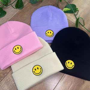 以下が含まれることがあります： ピンク、ラベンダー、クリーム、黒の4色のニット帽。各帽子には、折り返されたつばに黄色のスマイリーフェイスパッチが付いています。帽子は柔らかいニット素材で作られています。