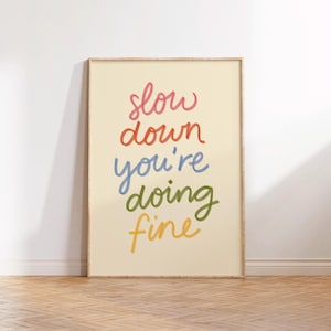 May include: A framed art print featuring the phrase "slow down you're doing fine" in a colourful, handwritten font. The text is arranged vertically on a cream background, enclosed within a light wood frame.