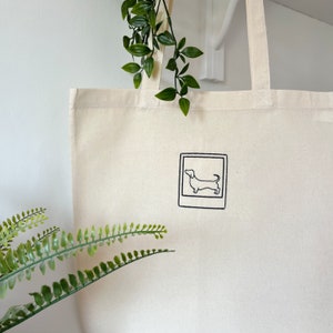 May include: A white canvas tote bag with a black outline of a dachshund dog inside a square. The bag has two long straps.