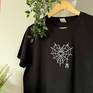 Puede incluir: Camiseta negra con un diseño de corazón de telaraña blanca y una pequeña araña colgando de la parte inferior.